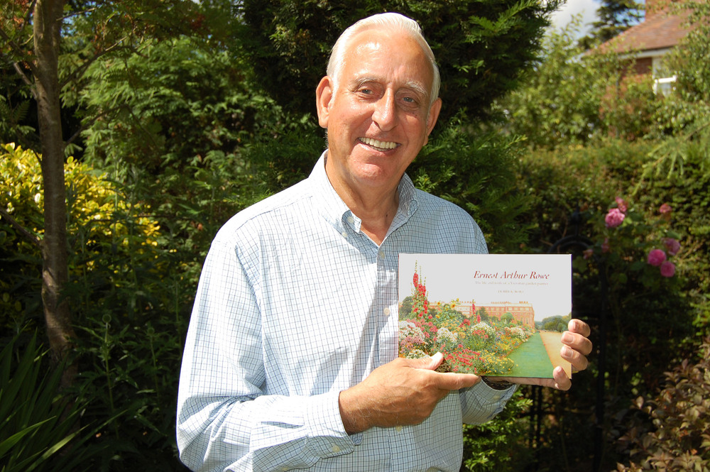 An gentleman with white hair standing in a garden and holding a copy of his family biography